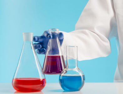 ręka chemika w fartuchu i rękawiczce stawiająca na stole zlewkę z substancją chemiczną